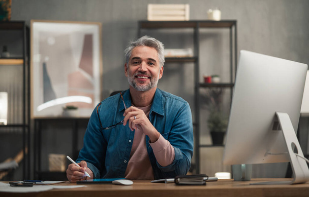 Ευτυχισμένος ώριμος άνθρωπος αρχιτέκτονας που εργάζεται σε tablet στο γραφείο σε εσωτερικούς χώρους στο γραφείο, κοιτάζοντας κάμερα. - Φωτογραφία, εικόνα