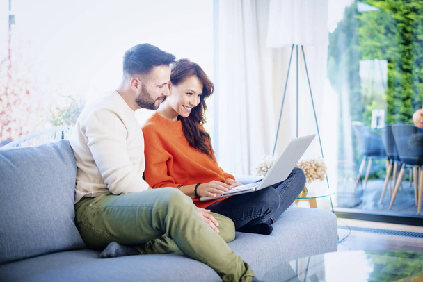 Happy couple relaxing on the couch together. Cheerful woman and handsome man using laptop while browsing on the internet. - Photo, Image
