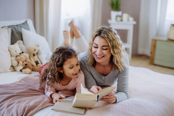 Happy mother with her little daughter lying on bed and reading book at home. - Foto, immagini