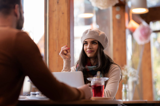 Candid shot of beautiful woman wearing beret hat and scarf while sitting at table with an unrecognizable man and drinking tea together at the cafe. - Foto, immagini