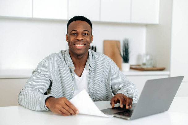 Positiv angenehmer afrikanisch-amerikanischer Typ, Student oder Freiberufler, in stylischer Freizeitkleidung, sitzt zu Hause mit Laptop in der Küche, schaut in die Kamera, lächelt freundlich - Foto, Bild