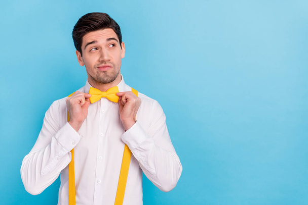 Photo of charming thoughtful guy dressed white shirt correcting yellow bowtie looking empty space isolated blue color background - Фото, зображення