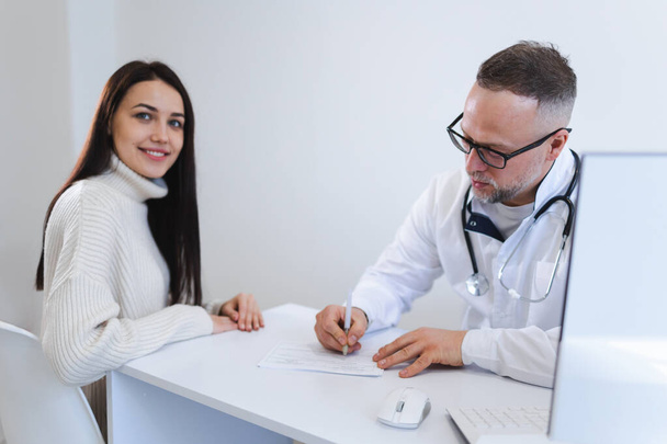 Il medico maschio scrive una prescrizione per la paziente femminile. Donna nello studio medico durante la consultazione - Foto, immagini