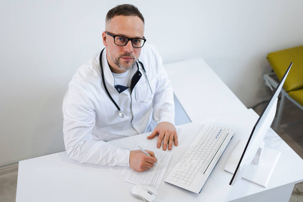 Médico varón llenando documentación médica mientras está sentado en su escritorio en la oficina - Foto, Imagen