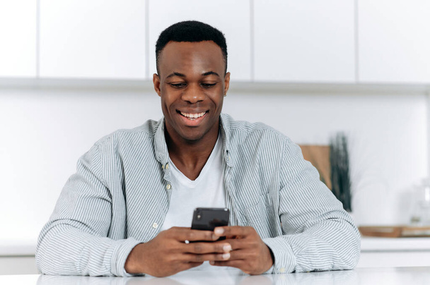 African American attractive positive young man, in stylish casual clothes, spend leisure time sitting at the table in the kitchen uses smartphone, browses internet, chatting in social media, smiles - Foto, Imagem