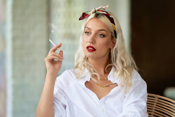 Portrait of beautiful green eyed young woman wearing hair scarf and white shirt while smoking cigarette. - Foto, Bild