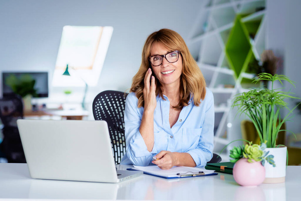 Glückliche Geschäftsfrau sitzt hinter ihrem Computer und telefoniert während der Arbeit.  - Foto, Bild