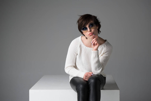 Opgewonden jonge vrouw met een zonnebril zittend op witte kubus in studio geïsoleerd over grijze achtergrond, kijkend naar camera. Trendy meid met verrassende gezichtsuitdrukking. - Foto, afbeelding