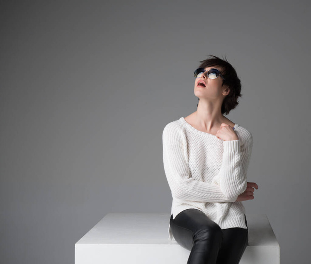 Excited young woman with sunglasses sitting on white cube in studio isolated over gray background, looking at camera. Trendy girl with surprised face expression. - Foto, Imagem