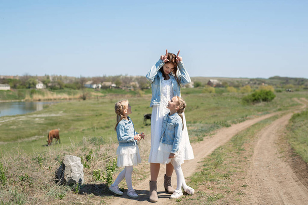 Mother with her twin daughters walks along a dirt road in a village near cows. Mother and daughters pretend to be cows and have fun - Foto, Imagem