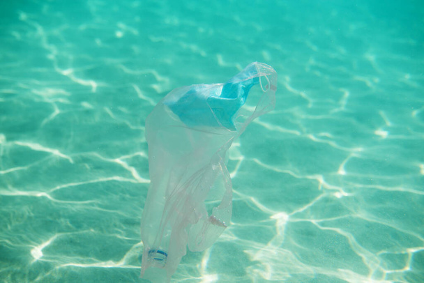 Ocean pollution. Personal protective mask, plastic bottle and bag underwater. Consequences of overuse of surgical masks and improper waste disposal during the coronavirus pandemic. - Фото, изображение