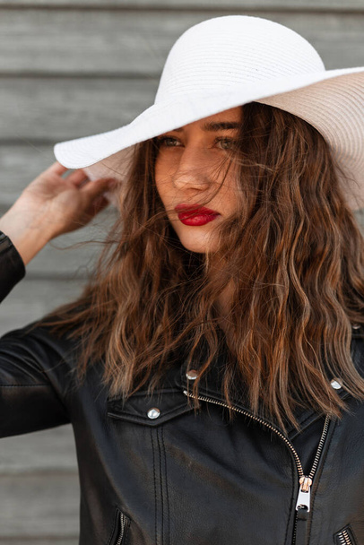 Portret mooie jonge vrouw met sexy rode lippen in zwarte mode leren jas in witte trendy zomerhoed in de buurt van houten muur op straat. Aantrekkelijk vrij stijlvol meisje rechtzet elegante hoed. - Foto, afbeelding