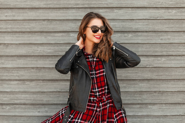 Lovely pretty young joyful woman with pretty smile in fashion youth red-black clothes in sunglasses posing near wooden vintage wall on street. Fashion model cute happy smiling woman rests in city. - Foto, Imagem
