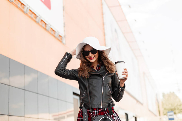 Mujer sonriente con estilo urbano en ropa de moda rojo-negro en gafas de sol en sombrero blanco con sabroso té en la ciudad. Chica de moda con labios rojos sexy y linda sonrisa con café en traje de moda camina al aire libre. - Foto, Imagen