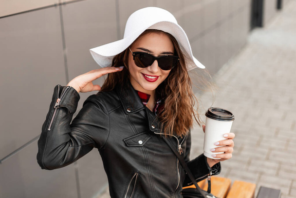 Πορτρέτο χαμογελαστή γυναίκα με φλιτζάνι τσάι ή καφέ με κόκκινα χείλη σε κομψό καπέλο σε μαύρο δερμάτινο σακάκι μόδας γυαλιά ηλίου κοντά σε σκούρο τοίχο στο δρόμο. Ευτυχισμένη χαριτωμένη γυναίκα με χαμόγελο κάθεται στον πάγκο στην πόλη. - Φωτογραφία, εικόνα