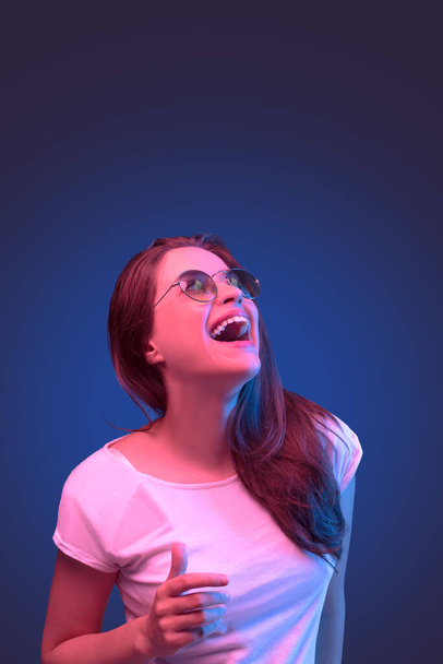 Néon gros plan portrait de jeune femme lunettes de soleil rondes et robe blanche sur fond bleu foncé - Photo, image