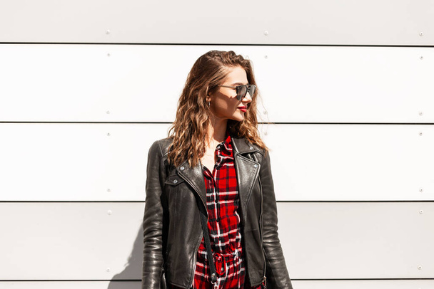 Kaunis hipster tyttö muodikas vaatteita aurinkolasit, nahkatakki ja laukku lähellä valkoista seinää kaupungissa - Valokuva, kuva