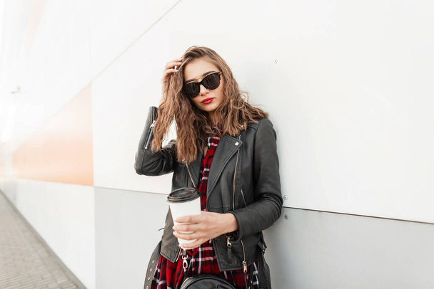 Fashion portrait woman in trendy red-black youth clothes from new collection with cup tea or coffee rest near modern wall on street. Gorgeous vogue beautiful woman with smile and hot drink outdoors. - Фото, изображение