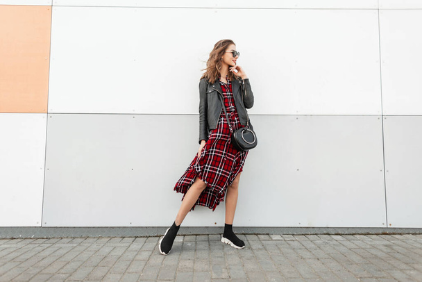 Boldog csinos nő piros-fekete alkalmi ruhák divatos napszemüveg divatos tornacipő vintage táska állványok és mosolyog a modern fal közelében fényes nappal. Mosolygó lány pózol az utcán a városban. - Fotó, kép
