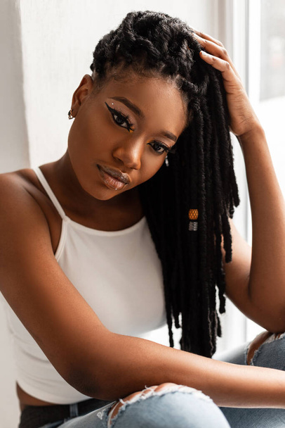 Beautiful young African American woman in fashionable white casual youth stylish white T-shirt near window in room. Fresh beauty portrait trendy black girl model with beauty makeup and dreadlocks. - Φωτογραφία, εικόνα