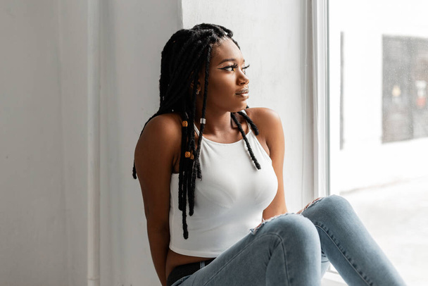 Mulher afro-americana bonita na moda top branco em jeans azul elegante com dreadlocks legal penteado relaxa na sala de janela perto. Menina preta legal na moda roupas jeans de verão - Foto, Imagem