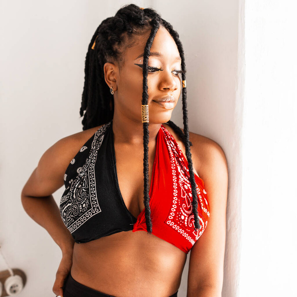 Portrait American black woman with trendy dreadlock hairstyle in fashionable youth swag red-black bandana top near white vintage wall in studio. Attractive afro girl model looking out window. - Photo, Image