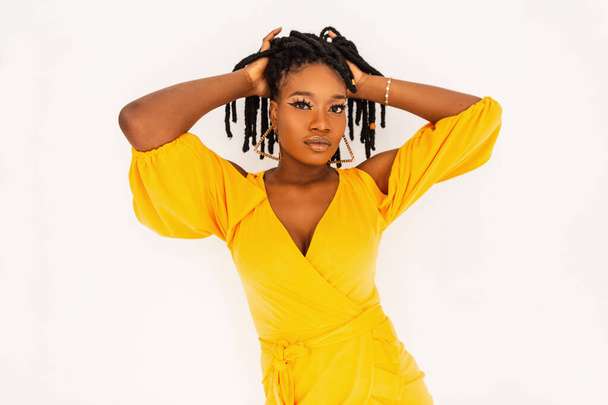 Docela elegantní afro žena v krásné módní letní žluté šaty narovnává dredy na bílém pozadí ve studiu. Krásná africká dívka v barevné elegantní oblečení z nové kolekce uvnitř. - Fotografie, Obrázek