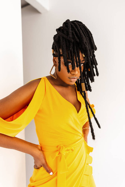 Gorgeous young African woman with cool dreadlocks in fashionable yellow dress stands near white wall in studio. Attractive black girl model in summer colored elegant outfit. Hairstyle cover face - Photo, Image