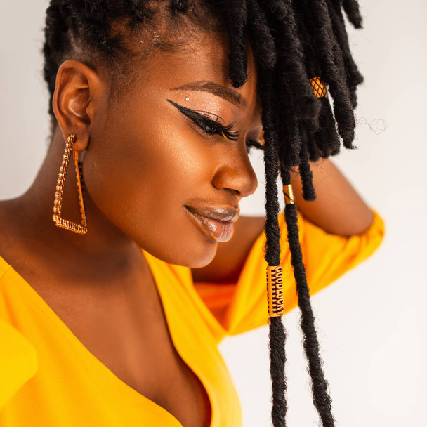 Fashion portrait of beautiful happy african girl with long dreadlocks hairstyle in fashionable yellow dress with gold earrings - Foto, Bild