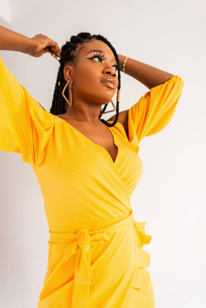 Glamorosa mujer afro en hermoso vestido amarillo de verano de moda endereza rastas sobre fondo blanco en studio.Sexy chica afroamericana en traje elegante colorido de la nueva colección en la habitación - Foto, imagen