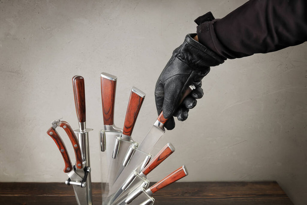 Χέρι στο μαύρο δερμάτινο γάντι παίρνει μαχαίρι κουζίνας από το σύνολο στο τραπέζι, έννοια της επίθεσης στο σπίτι και ποινική, κοντινό πλάνο - Φωτογραφία, εικόνα