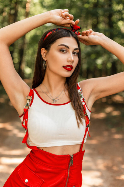 Sexy attractive young woman with red lipstick in vintage bandana in fashionable summer red-white clothes posing outdoors on sunny day. Beauty portrait of fashionable glamorous girl in park. Youth look - Фото, изображение