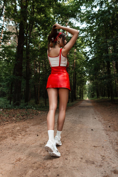 Πλάτη άποψη ενός μοντέρνου κοριτσιού με όμορφα πόδια σε μια κομψή κόκκινη φούστα σε λευκό top σε vintage sneakers στο δάσος. - Φωτογραφία, εικόνα