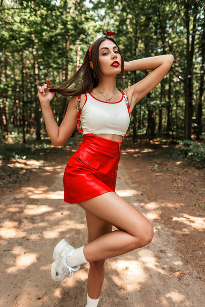 Pretty girl with red lips with sexy legs in a red fashionable short dress in sneakers walks in a park on summer  holidays. - Фото, зображення