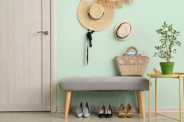 Ψάθινα καπέλα, πάγκος, παπούτσια και houseplant κοντά στο χρώμα τοίχο στο διάδρομο - Φωτογραφία, εικόνα