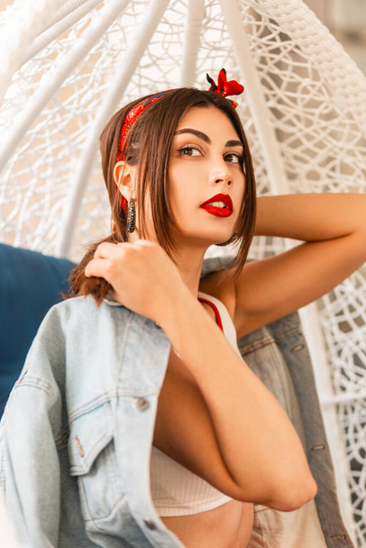 Mulher de moda bonita com lábios bonitos vermelhos com uma bandana elegante em uma jaqueta de jeans em um top branco elegante posa em uma cadeira de ovo vintage ao ar livre. - Foto, Imagem