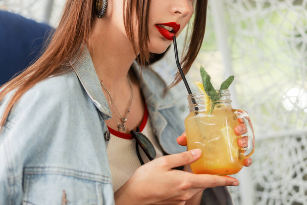Cute woman in fashion denim jacket with red lipstick and sexy lips drinks sweet fruity summer lemonade with ice, mint and fresh lemon slices on terrace in cafe. Close-up. - Photo, Image