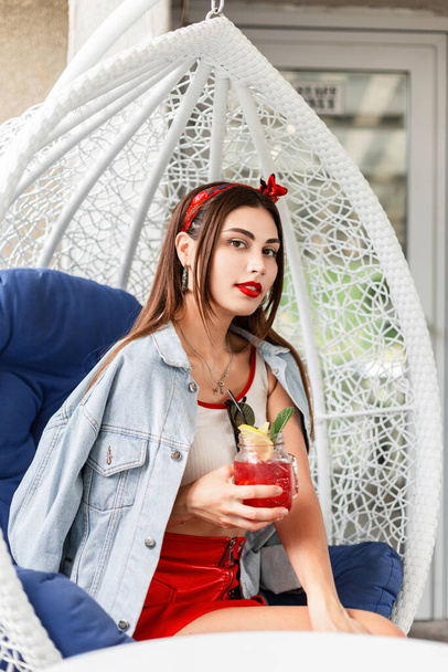 Fashionable young woman in beauty summer clothes with delicious berry-fruit lemonade sits on vintage armchair on terrace. Lovely girl resting and enjoys sweet citrus drink with ice and mint in cafe. - Foto, imagen