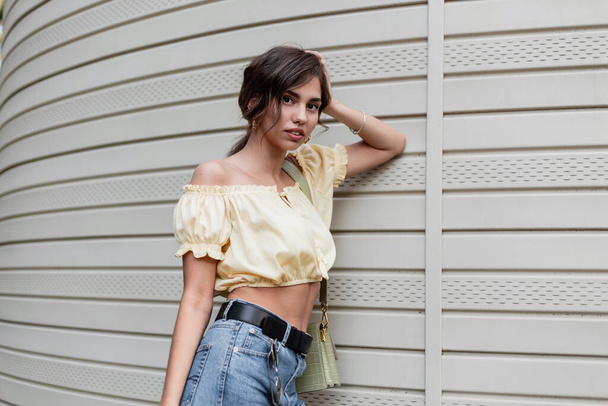 Pretty young girl in a yellow blouse with blue jeans and a fashionable handbag stands near the siding wall - Photo, Image