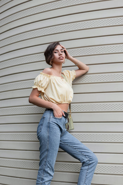 Fashionable pretty woman hipster in a summer yellow stylish blouse and vintage blue high waist jeans with a handbag stands near the siding wall - Photo, Image