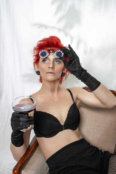 Portrait of a European woman with bright red hair in a black bra and sunglasses in her hand adorned with pearls and rhinestones and a glass of red wine - Fotoğraf, Görsel