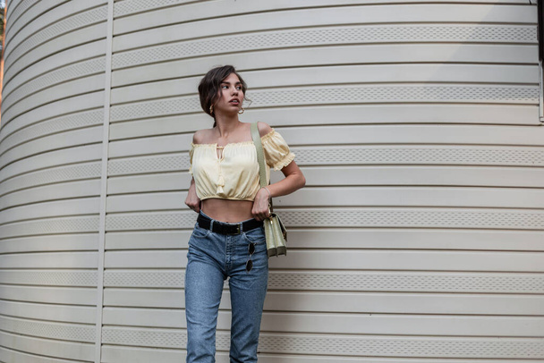 Fashion hipster beautiful trendy curly girl in a fashionable casual outfit with yellow summer top blouse, blue jeans and  leather bag stands near a siding wall - Foto, imagen