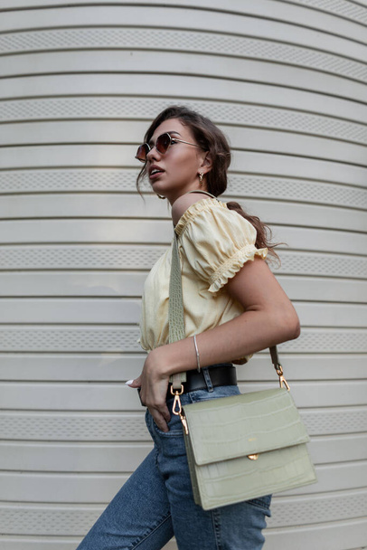 Mode hipster femme bouclée dans des lunettes de soleil rondes vintage dans un haut de chemisier jaune en jean bleu élégant avec un sac à main marche près du mur de revêtement de la maison - Photo, image