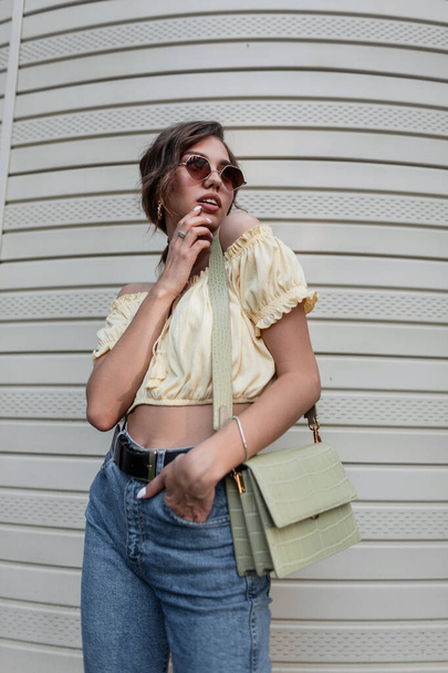 Piuttosto alla moda donna hipster con occhiali da sole e borsa in classico jeans blu in posa vicino al muro - Foto, immagini