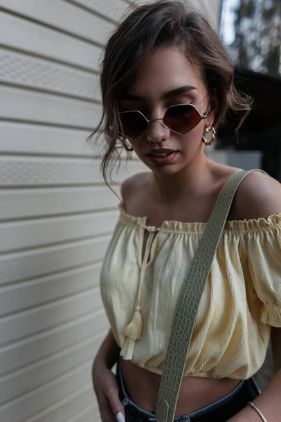 Portrait cool de femme à la mode avec des lunettes de soleil rondes élégantes en chemisier jaune marche près du mur sur la rue - Photo, image