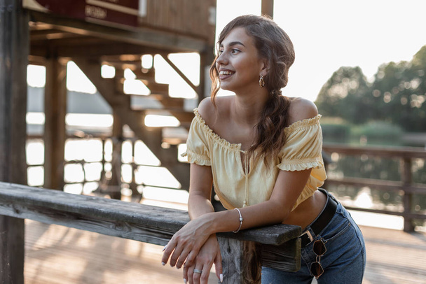 Довольно счастливая девушка с улыбкой в модной джинсовой одежде с летним желтым топом стоит на деревянном пирсе у озера - Фото, изображение