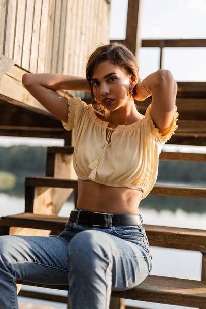 Young beautiful woman hipster in a yellow blouse top and blue jeans sits on wooden steps outdoors - Photo, Image