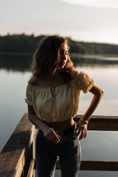 Pretty hipster woman with curly hair in blue fashionable jeans with yellow blouse walks in nature near the lake at sunset - Фото, изображение