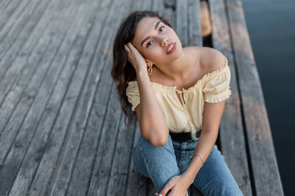 Pretty young woman in a fashionable yellow blouse with blue jeans sits on a wooden pier - Foto, immagini