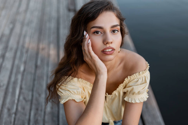 Frisches Sommerporträt einer hübschen Frau in modischem gelben Blusenoberteil sitzt auf einem Holzsteg am See und blickt in die Kamera.. - Foto, Bild
